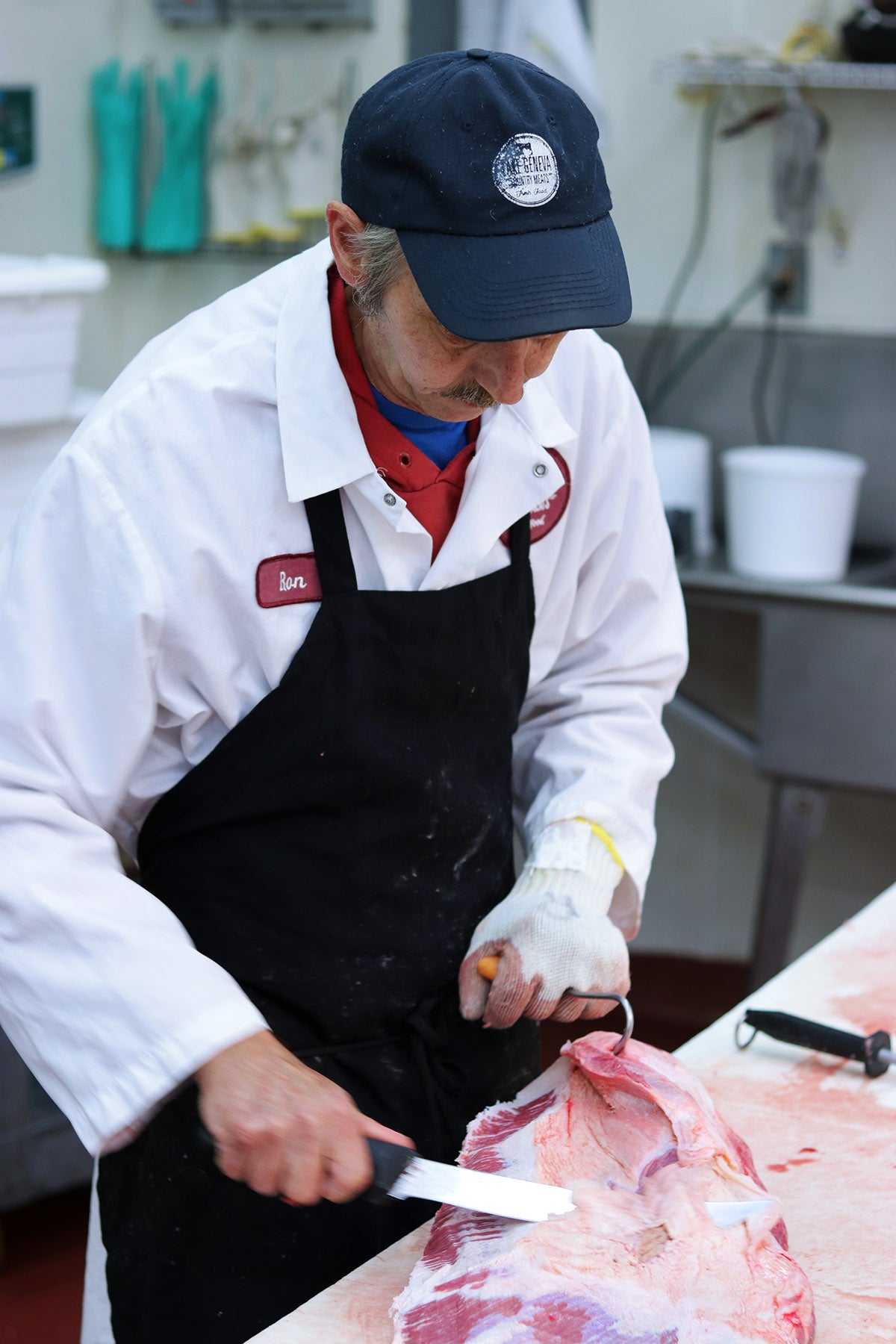 Butcher in a white coat and black apron cutting meat with a knife in a kitchen setting.