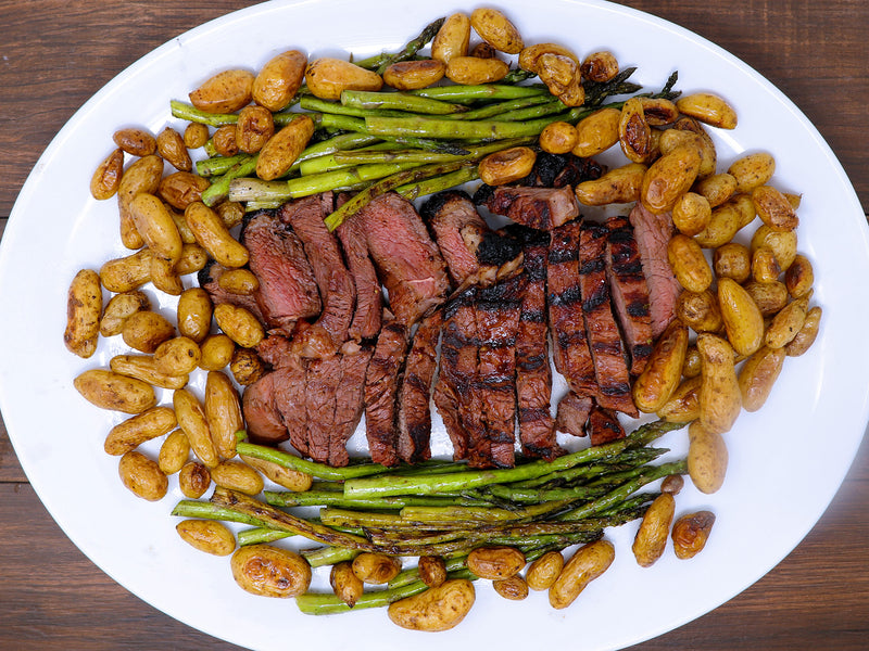 A top-down view of a plated meal featuring sliced grilled steak on a large white plate, surrounded by roasted potatoes and green asparagus on a dark wooden table.