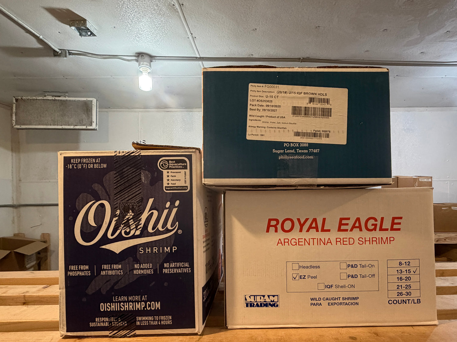 Three cardboard boxes labeled 'Oishii Shrimp' and 'Royal Eagle Argentina Red Shrimp' stacked on a wooden floor.