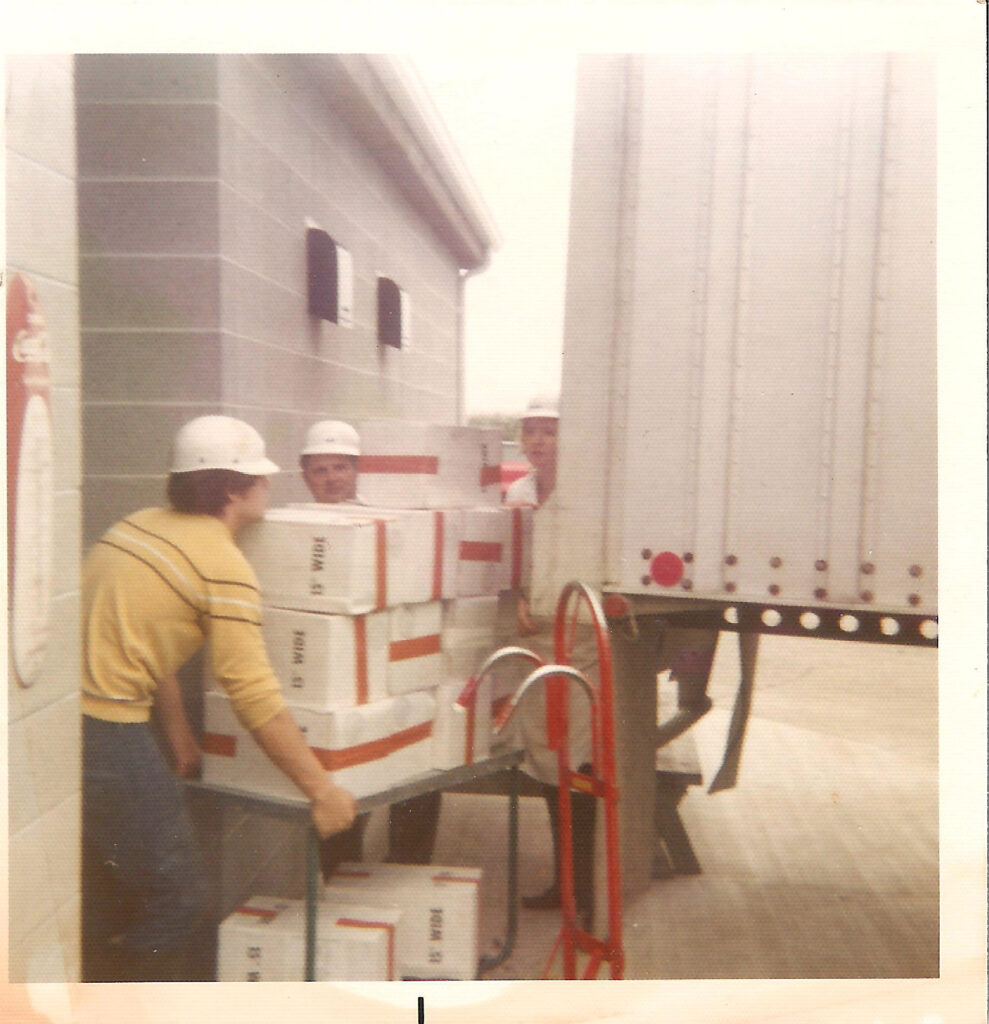 John Leahy (peaking over the boxes) supervising employees unloading a semi full of butcher paper. 